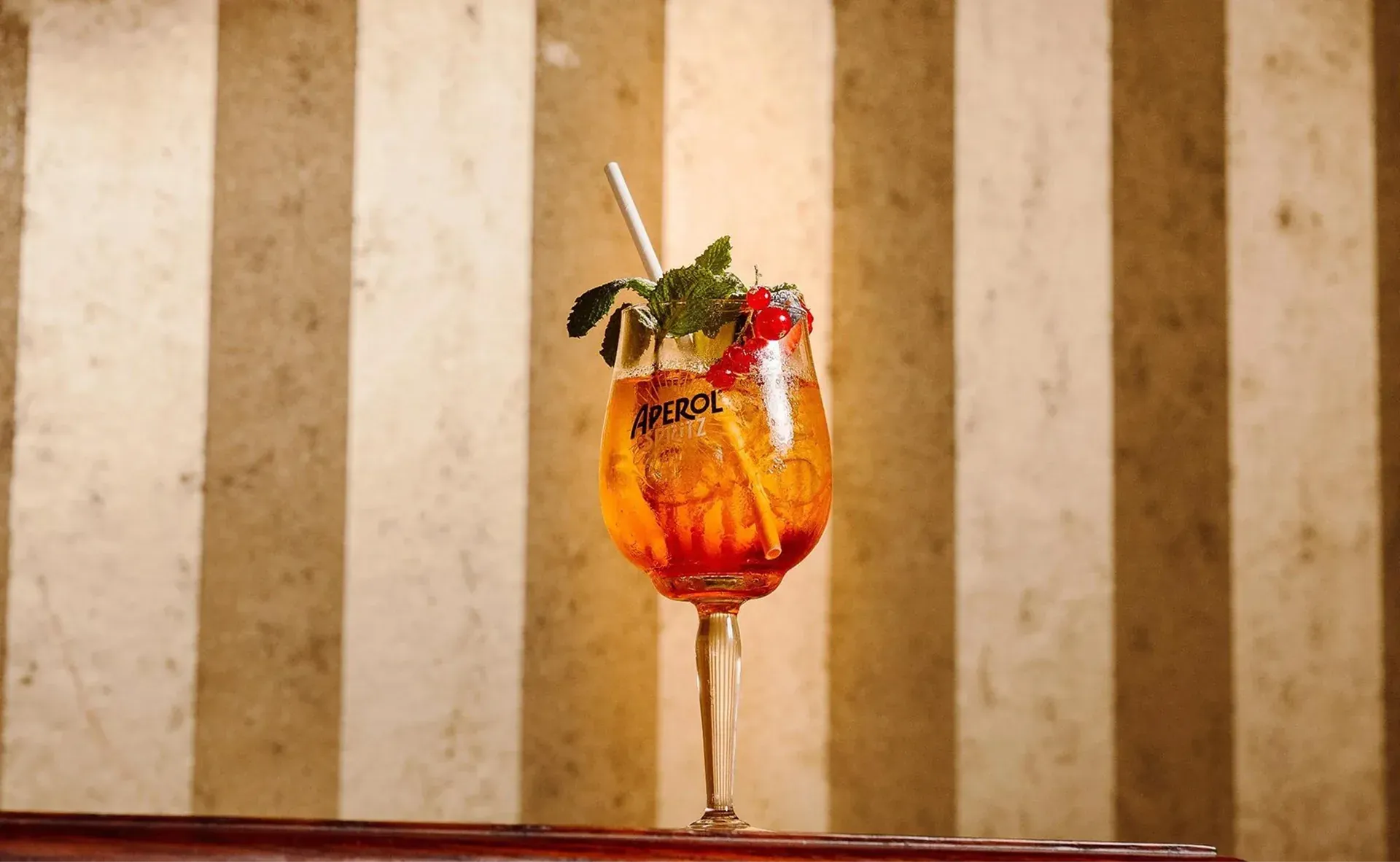 An Aperol Spritz garnished with mint and redcurrants against a striped wall