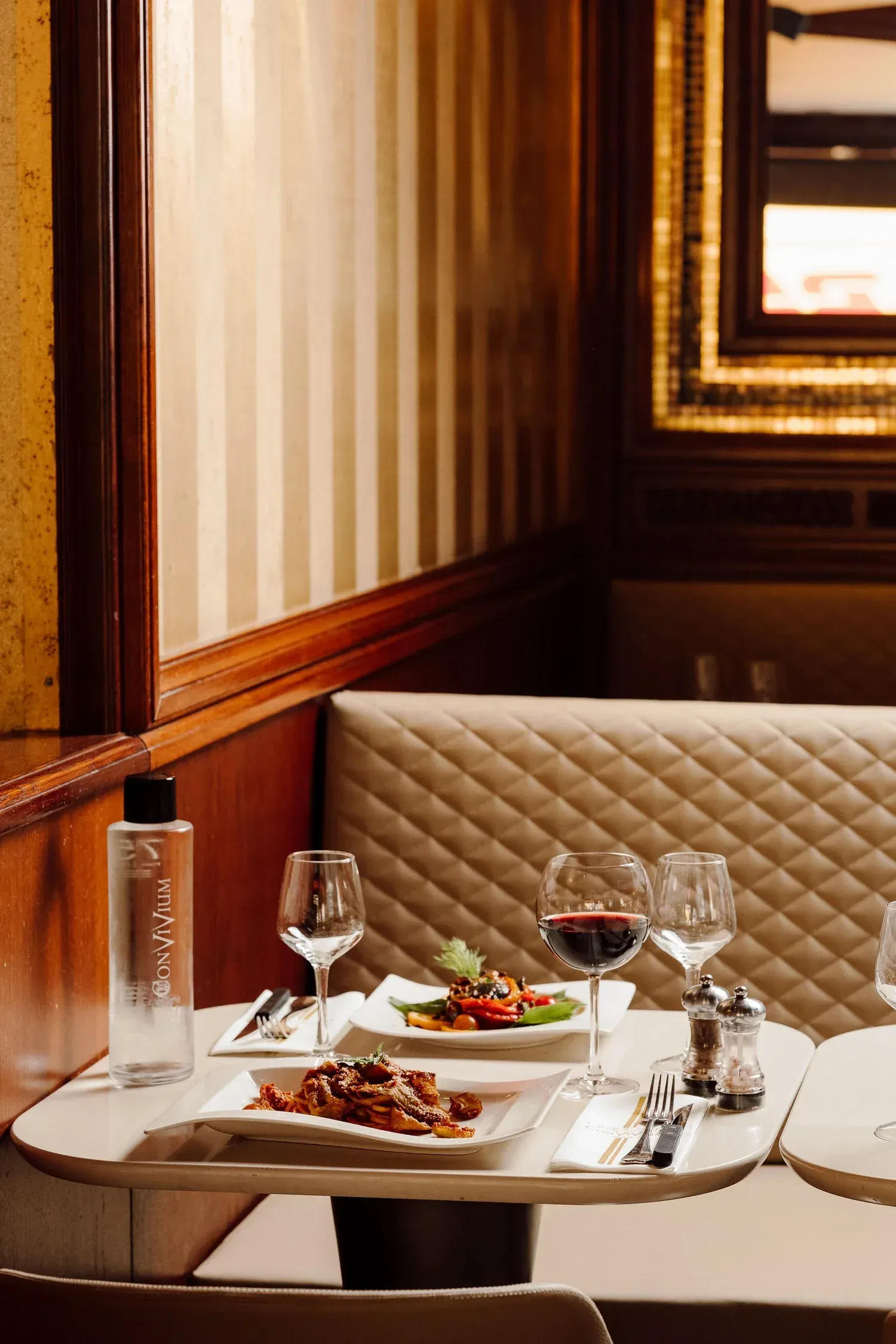 Table set for two in a leather banquette, with pasta, a glass of red wine and a branded Convivium water bottle