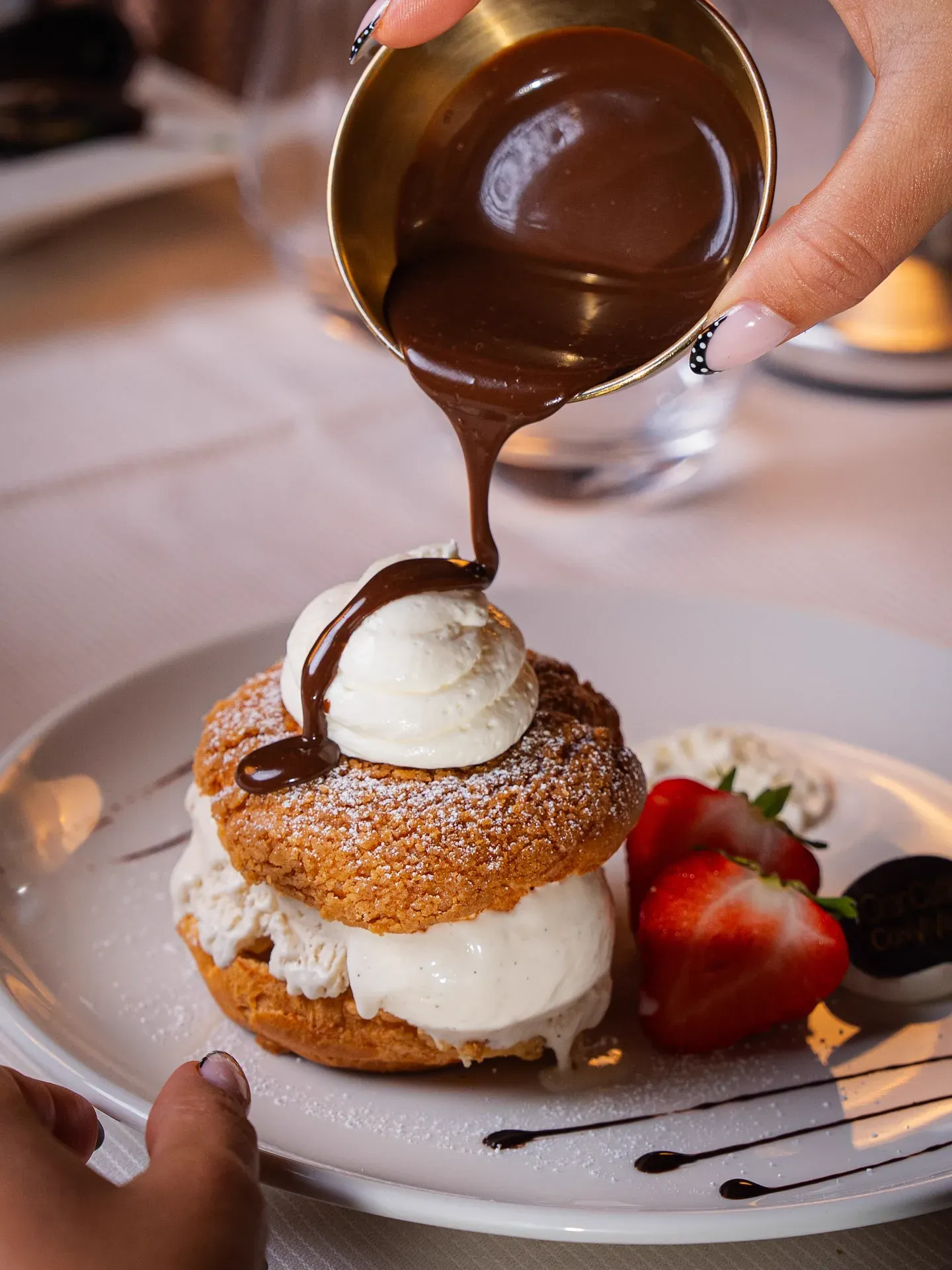 Warm chocolate sauce poured over a profiterole with whipped cream and strawberries