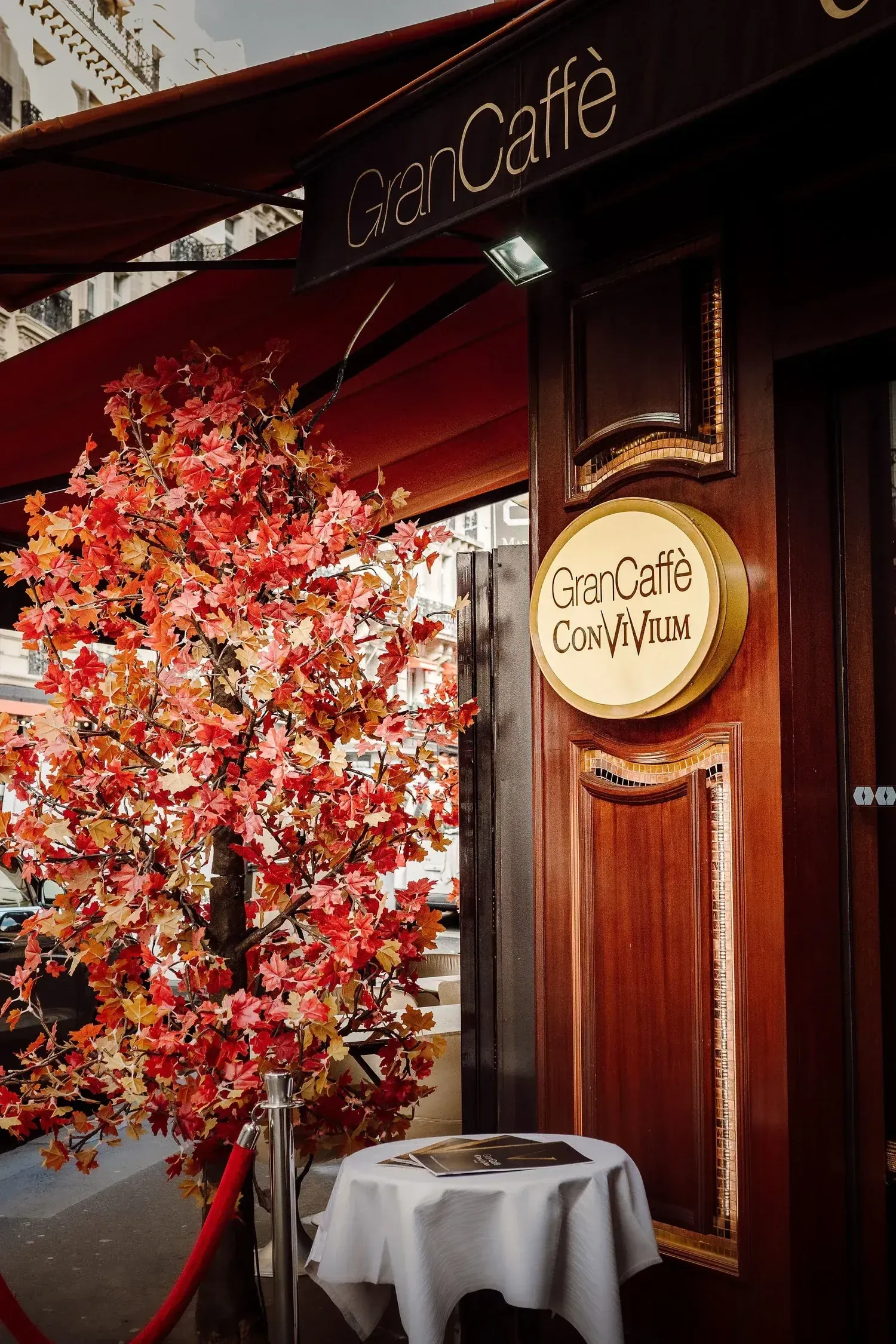 Gilded Gran Caffè Convivium plaque beside the mahogany entrance, framed by an autumn tree on the terrace