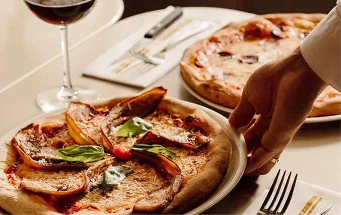 Two pizzas and a glass of red wine set on the table as a waiter reaches in