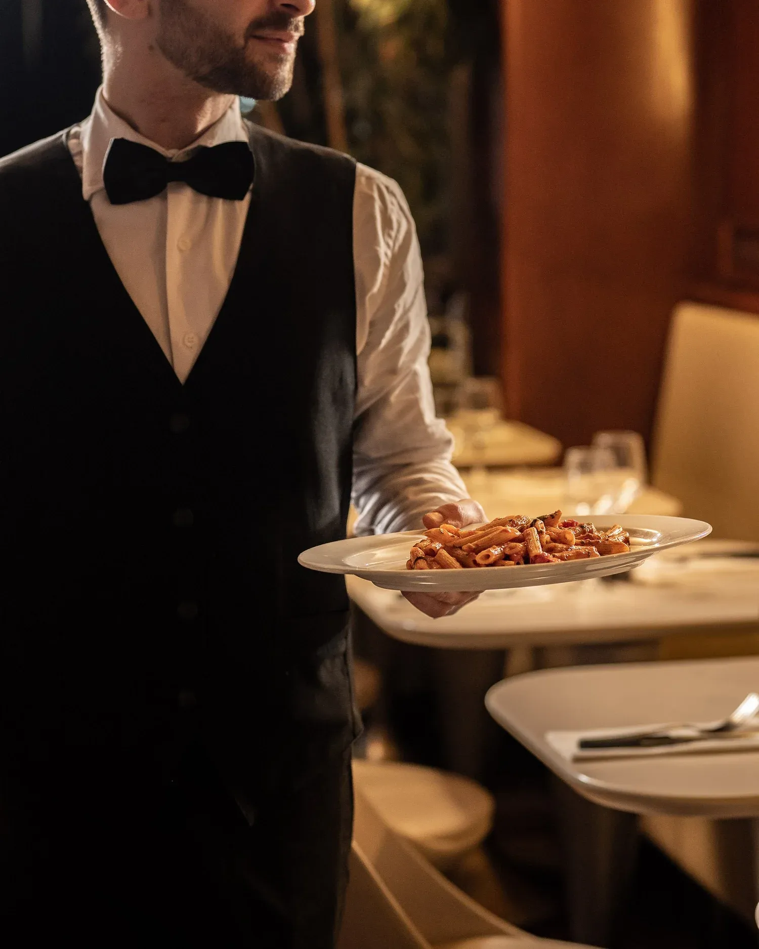 Waiter in waistcoat and bow tie carrying a plate of penne to a table